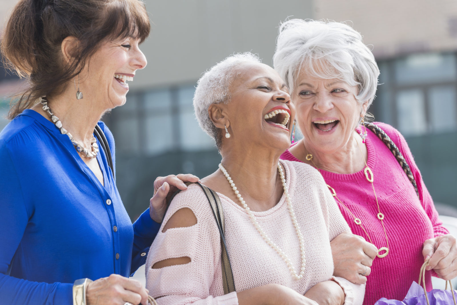 A group of three multi-ethnic senior women in their 60s and 70s standing outdoors, laughing.