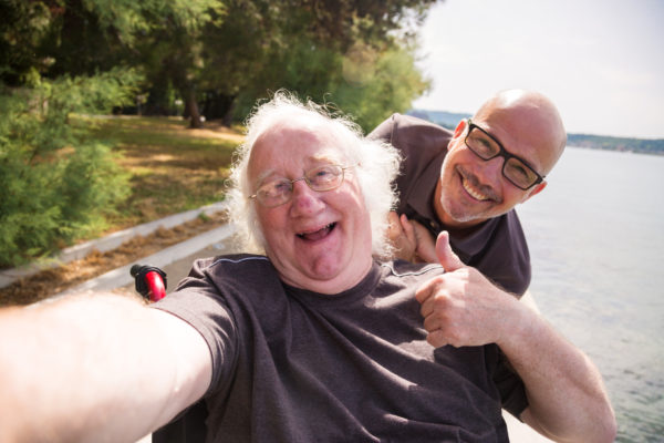 old man in wheelchair taking a selfie with his phone with caregiver in background