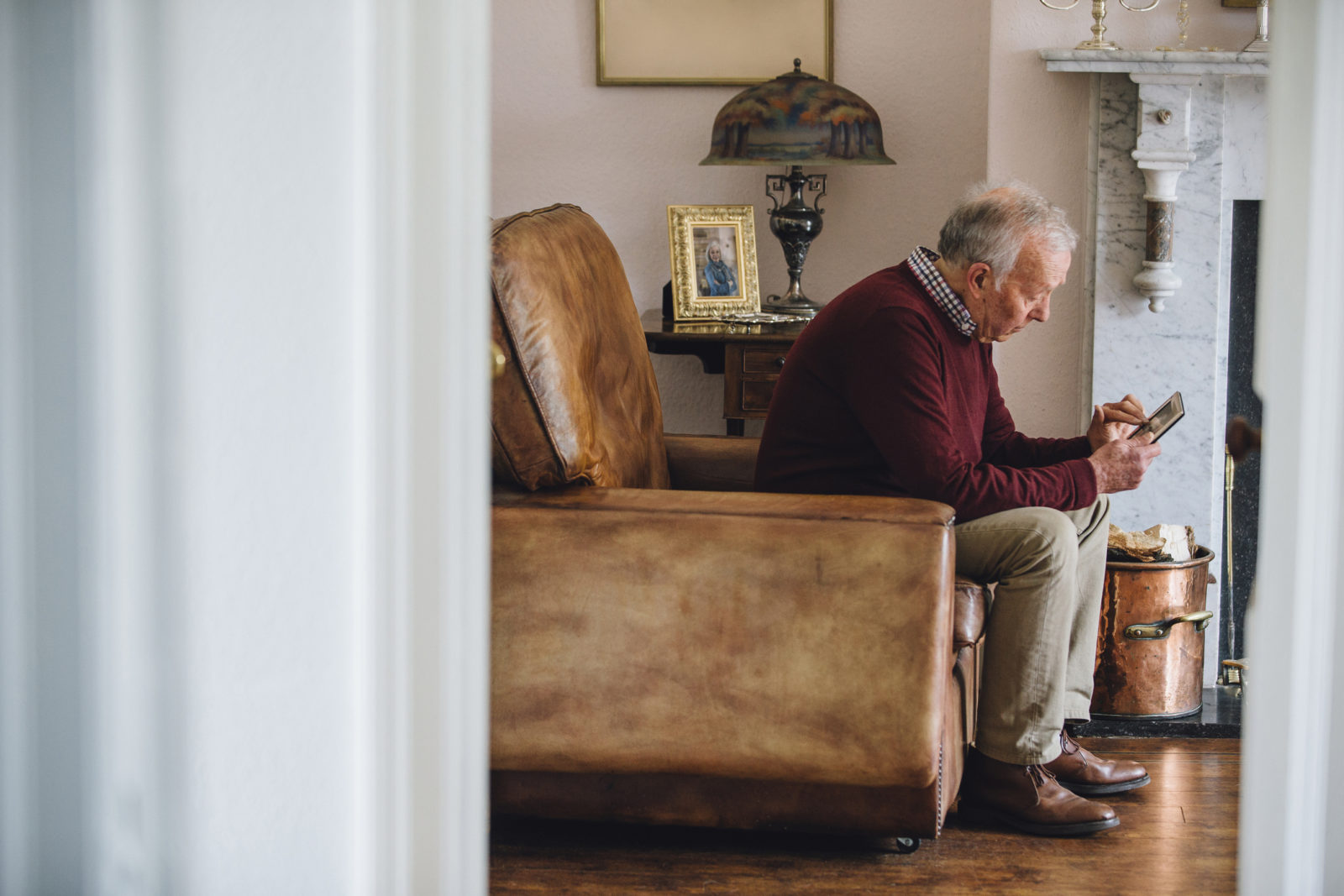 Senior man is sitting in an armchair in the living room of his home, holding and looking at an old photo with a sad expression.