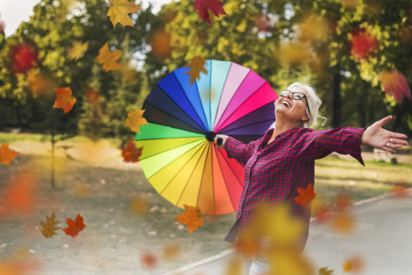 Woman with multi-colored parasol walking through falling autumn leaves in park
